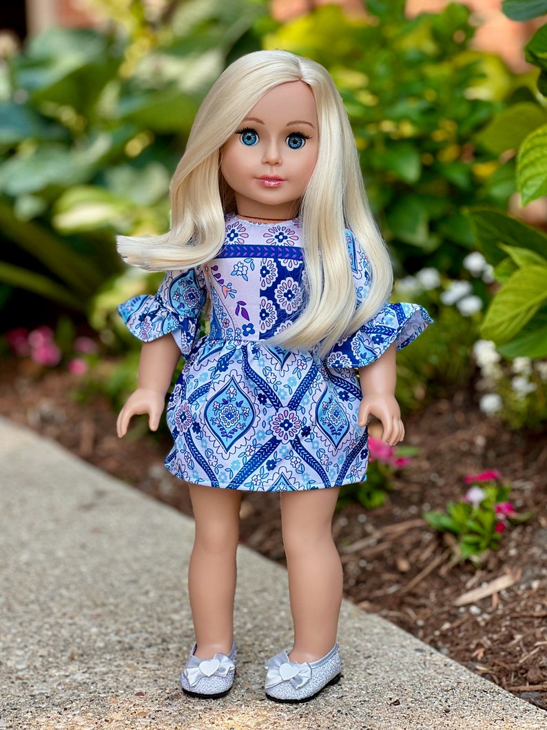 May include: A blonde doll wearing a blue and white patterned dress with silver shoes. The doll has blue eyes and is standing on a gray sidewalk.
