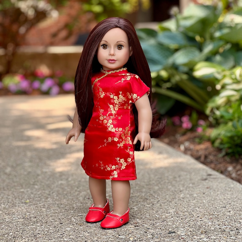 May include: A red and gold Chinese-style dress with floral patterns, worn by a doll with long brown hair and brown eyes. The doll is wearing red shoes and standing on a gray sidewalk.