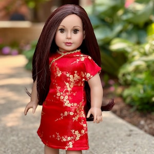 May include: A red and gold doll dress with a floral pattern. The doll is wearing a red dress with gold trim and a floral pattern. The dress has a mandarin collar and short sleeves.