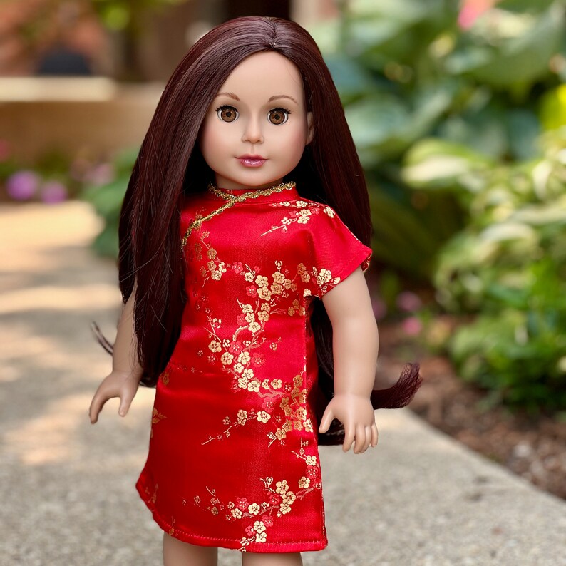 May include: A red and gold doll dress with a floral pattern. The doll is wearing a red dress with gold trim and a floral pattern. The dress has a mandarin collar and short sleeves.