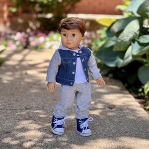 May include: A doll with brown hair and blue eyes wearing a white t-shirt with thin horizontal stripes, a blue denim jacket with grey jersey sleeves and a hood, grey sweatpants, and blue high-top sneakers with white laces and white rubber toes.