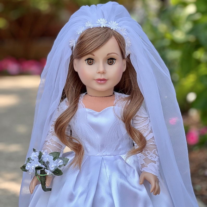 May include: A white satin dress with lace sleeves and a white tulle veil with a floral headband. The doll is wearing a white satin dress with lace sleeves and a white tulle veil with a floral headband.
