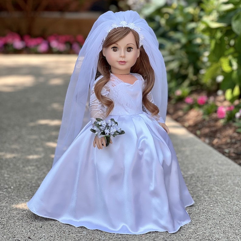 May include: A white satin wedding dress with lace sleeves and a tulle veil for a doll. The doll is holding a small bouquet of white flowers.