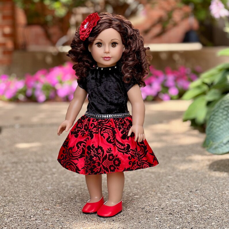 May include: A doll with long curly brown hair and a red flower hair accessory. The doll wears a black velvet top with a silver-studded neckline and a red skirt featuring a black damask pattern and a silver-studded waistband, paired with red shoes.