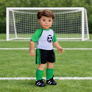 Puede incluir: Una muñeca de cabello castaño que viste una camiseta de fútbol blanca y verde con un gráfico de balón de fútbol en blanco y negro en el pecho, pantalones cortos negros con rayas laterales verdes, calcetines verdes y zapatillas deportivas.