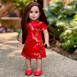May include: A doll wearing a red and gold cheongsam dress with a floral pattern. The doll has long brown hair and is wearing red shoes.