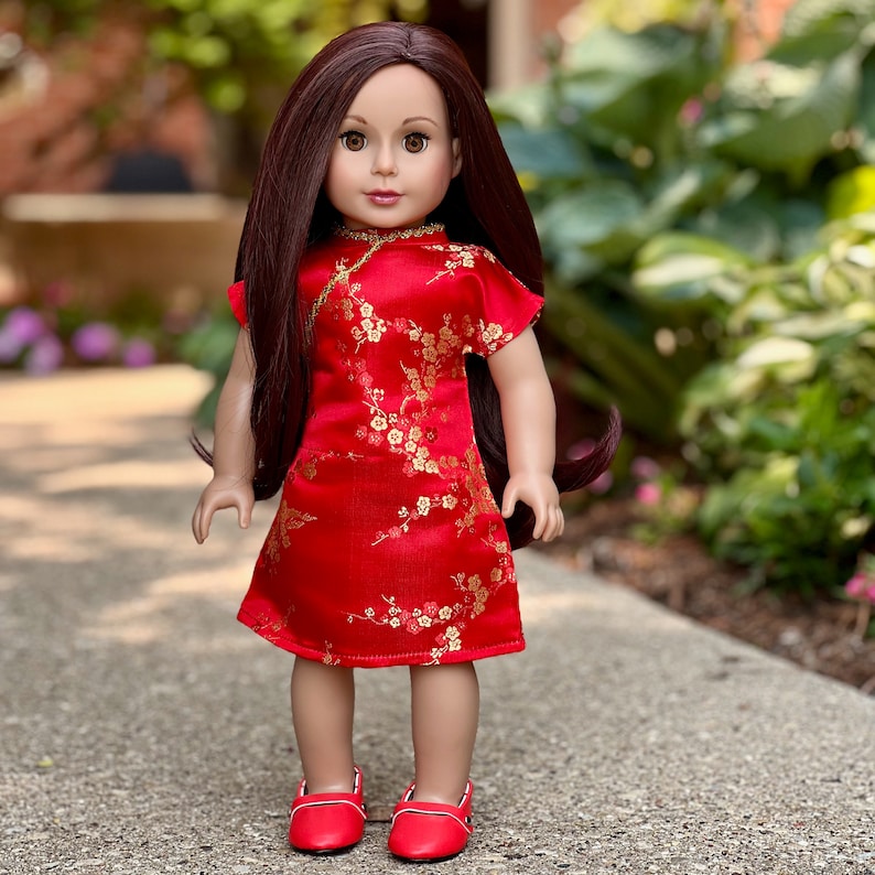 May include: A doll wearing a red and gold cheongsam dress with a floral pattern. The doll has long brown hair and is wearing red shoes.