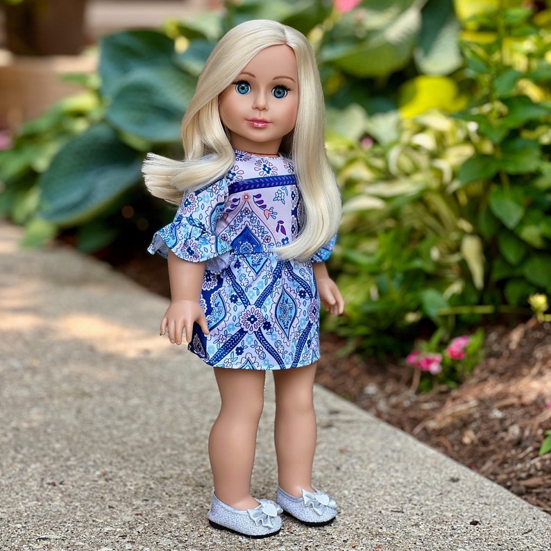 May include: A doll with long blonde hair wearing a blue and purple patterned dress with a pink and white floral design. The doll is wearing silver shoes with a flower design.