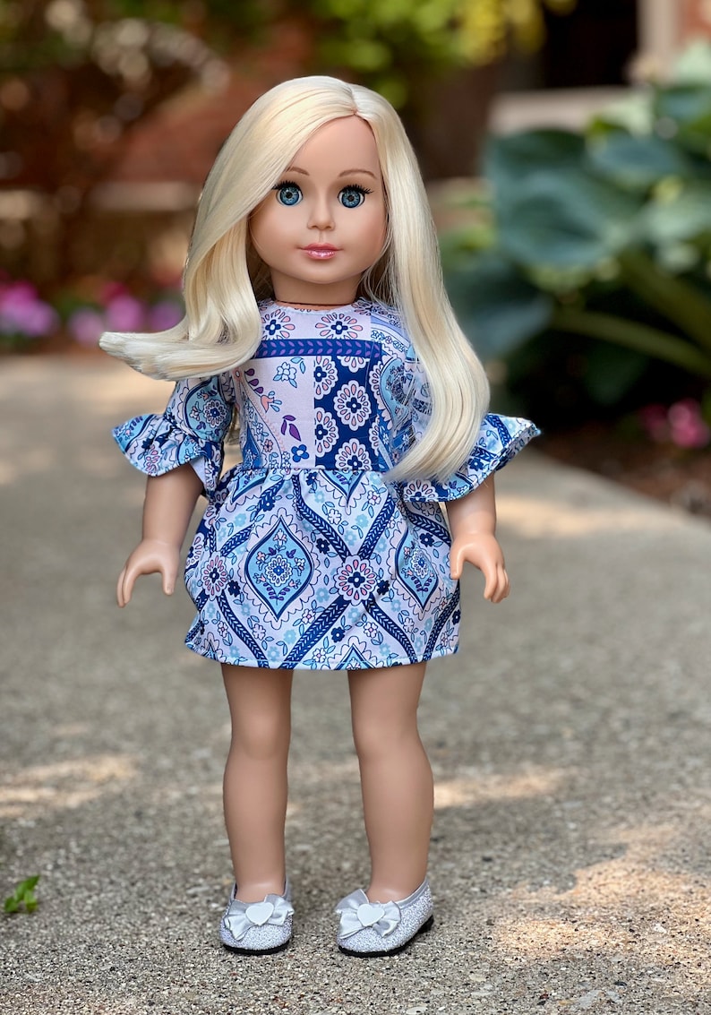 May include: A blonde doll wearing a blue and white patterned dress with ruffled sleeves and silver shoes.