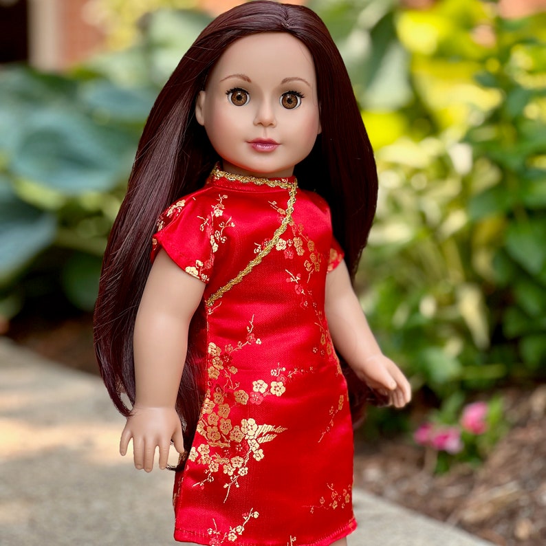 May include: A doll wearing a red and gold Chinese-style dress with floral patterns. The doll has long brown hair and brown eyes.