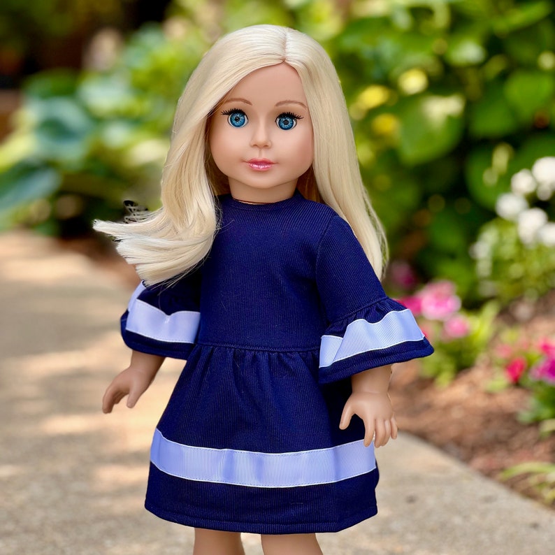 K&ouml;nnte beinhalten: Eine Puppe in einem marineblauen Kleid mit wei&szlig;en Verzierungen. Das Kleid hat Glocken&auml;rmel und einen R&uuml;schen-Saum. Die Puppe hat langes blondes Haar und blaue Augen.