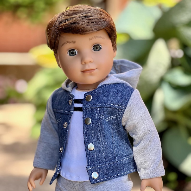 May include: A doll with brown hair and grey eyes wearing a white t-shirt with black stripes, a blue denim jacket with grey jersey sleeves and a hood, and grey pants. The jacket features silver-toned metal buttons down the front closure.