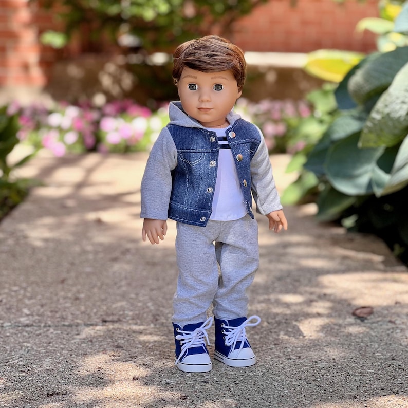 May include: A doll with brown hair and brown eyes wearing a white t-shirt, a blue denim jacket with gray jersey sleeves and a hood, gray sweatpants, and blue high-top sneakers with white laces and white rubber toe caps and soles.