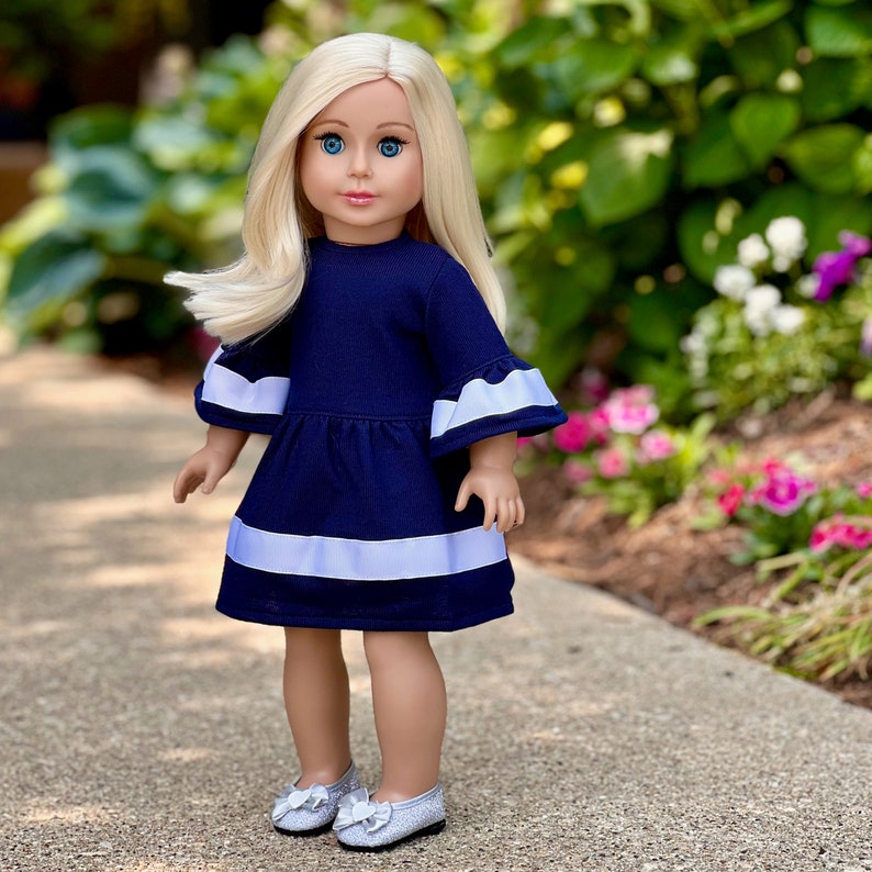 K&ouml;nnte beinhalten: Eine Puppe in einem dunkelblauen Kleid mit wei&szlig;em Besatz und silbernen Schuhen. Das Kleid hat einen R&uuml;schenkragen und R&uuml;schen&auml;rmel.