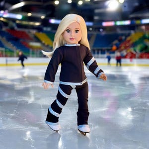 Könnte beinhalten: Eine Puppe mit blonden Haaren, die ein schwarzes Eiskunstlauf-Outfit mit silbernen Akzenten trägt. Die Puppe steht auf einer Eisbahn und trägt Eislaufschuhe. Das Outfit umfasst eine Jacke und eine Hose mit silbernen Streifen.