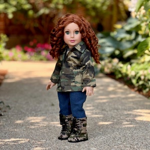 May include: A doll with long curly auburn hair and green eyes wearing a camouflage print jacket with front pockets, dark blue denim trousers, and matching camouflage print lace-up boots with black laces and silver eyelets on a light-coloured path.