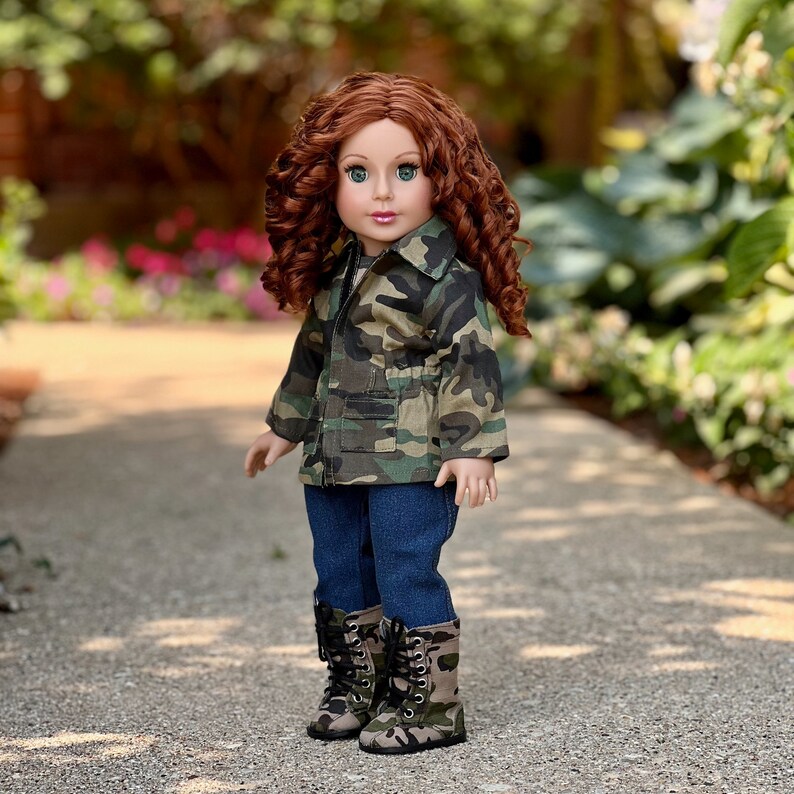 May include: A doll with long curly auburn hair and green eyes wearing a camouflage print jacket with front pockets, dark blue denim trousers, and matching camouflage print lace-up boots with black laces and silver eyelets on a light-coloured path.