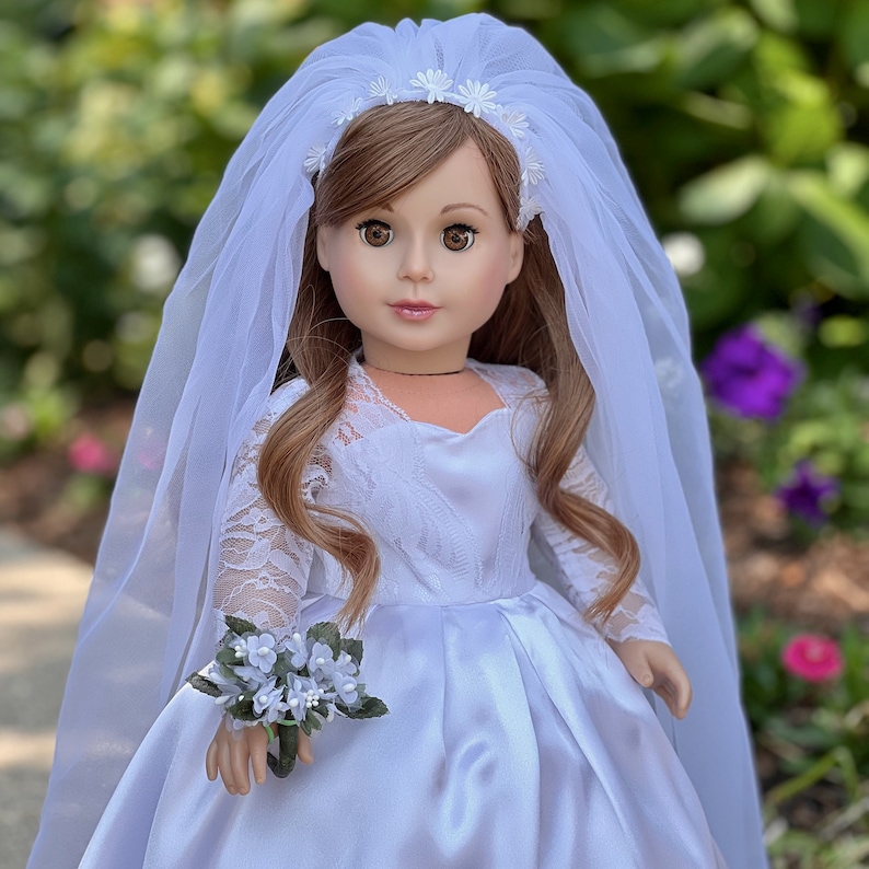 May include: A white satin bridal gown with lace sleeves and a white tulle veil with a floral headband. The doll is holding a bouquet of white flowers.