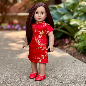May include: A doll wearing a red dress with gold floral embroidery and red shoes. The doll has long brown hair and brown eyes.
