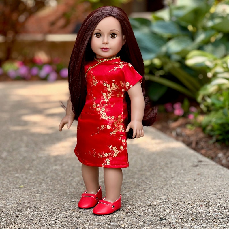 May include: A doll wearing a red dress with gold floral embroidery and red shoes. The doll has long brown hair and brown eyes.