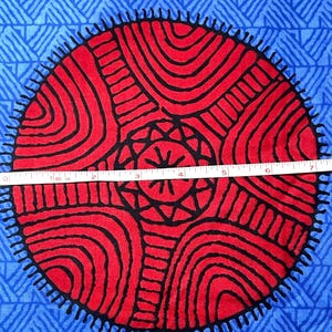 May include: Close-up of a fabric with a vibrant pattern. The design features large red circles with black curved lines and a central star-like shape, set against a blue background with a geometric pattern. A measuring tape shows the fabric is approximately 7 inches wide.