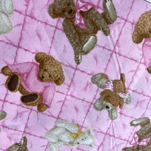 May include: Pink fabric with a quilted pattern featuring repeating illustrations of teddy bears. The bears are in various poses, some wearing pink sweaters or bows. The bears are brown and white.