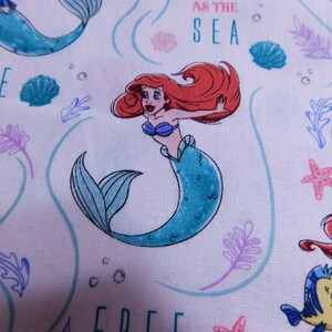 May include: White fabric with a repeating pattern of Ariel the Little Mermaid, seashells, seaweed, and the text "Free as the Sea".