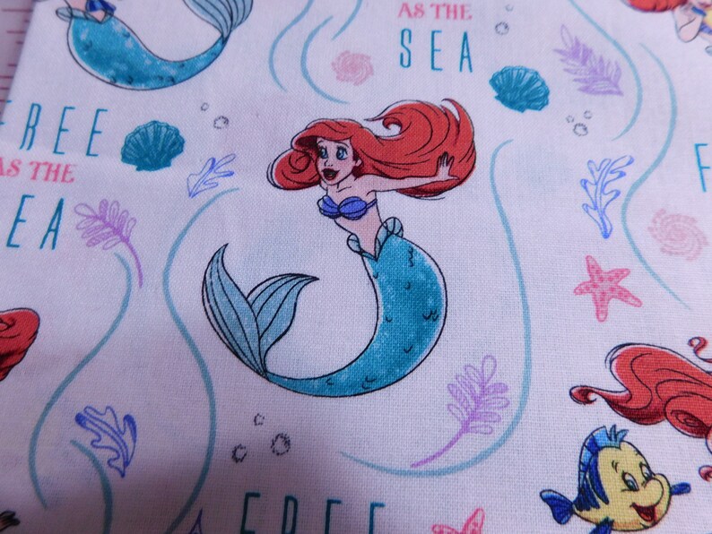 May include: White fabric with a repeating pattern of Ariel the Little Mermaid, seashells, seaweed, and the text "Free as the Sea".