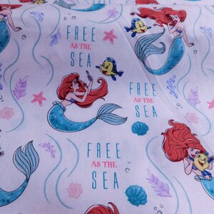 May include: White fabric with a repeating pattern of Ariel the Little Mermaid. She is depicted as a mermaid with red hair, a blue tail, and a seashell bra. The background is light blue with white swirls and stars. The text "Free as the Sea" is repeated throughout the fabric.