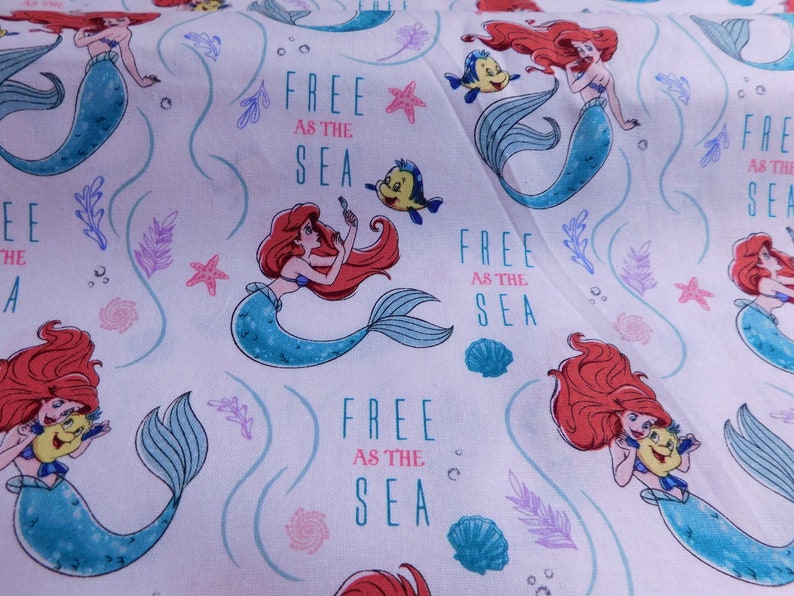 May include: White fabric with a repeating pattern of Ariel the Little Mermaid. She is depicted as a mermaid with red hair, a blue tail, and a seashell bra. The background is light blue with white swirls and stars. The text "Free as the Sea" is repeated throughout the fabric.
