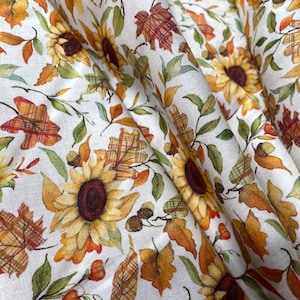 May include: White fabric with a repeating pattern of sunflowers, autumn leaves, and acorns in shades of yellow, orange, and brown. The fabric has a soft, textured appearance, perfect for fall-themed crafts and home decor projects.