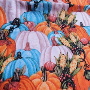May include: A fabric with a pattern of orange, white, and blue pumpkins. The pumpkins are arranged in a random pattern and are surrounded by green leaves and corn.