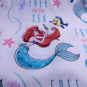 May include: A white fabric with a repeating pattern of Ariel the Little Mermaid, a yellow fish, and the text "Free as the Sea".
