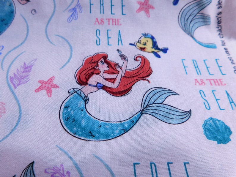 May include: A white fabric with a repeating pattern of Ariel the Little Mermaid, a yellow fish, and the text "Free as the Sea".