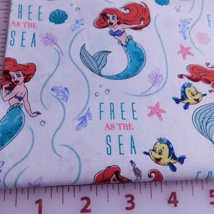 May include: White fabric with a repeating pattern of Ariel the Little Mermaid, Flounder, and the text "Free as the Sea".