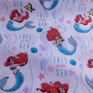 May include: White fabric with a repeating pattern of Ariel the Little Mermaid swimming with Flounder. The background is white with pink and purple seaweed and seashells. The text "Free as the Sea" is repeated throughout the fabric.