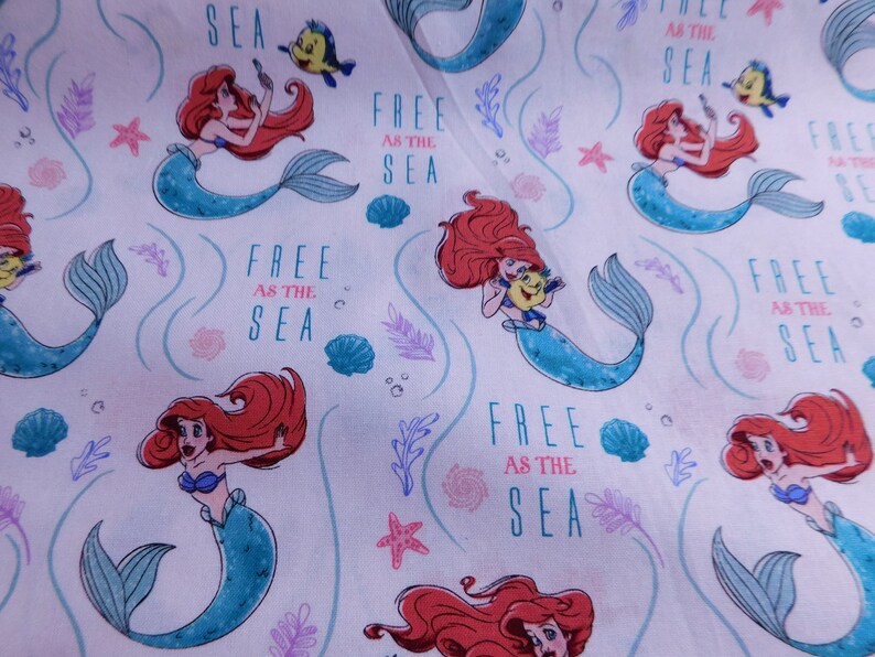 May include: White fabric with a repeating pattern of Ariel the Little Mermaid swimming with Flounder. The background is white with pink and purple seaweed and seashells. The text "Free as the Sea" is repeated throughout the fabric.