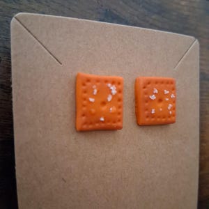 May include: A pair of orange square-shaped stud earrings, resembling crackers, with white salt details. The earrings are displayed on a brown earring card.