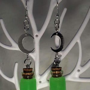May include: A pair of silver-toned earrings featuring a crescent moon design. Each earring has a small glass bottle filled with bright green liquid, topped with a cork. The earrings hang from silver-toned hooks.