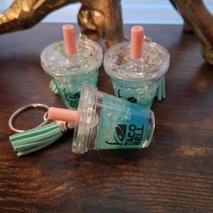 May include: Three miniature Taco Bell drink keychains. The clear plastic cups have a blue liquid and a pink straw. The Taco Bell logo is visible on the side of the cup. One keychain has a green and white tassel.