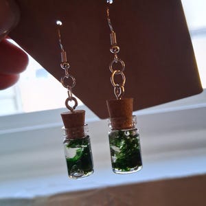 May include: A pair of handmade earrings featuring miniature glass bottles filled with green moss. Each bottle is sealed with a cork stopper and suspended from silver-colored ear wires. The earrings are displayed against a brown background.