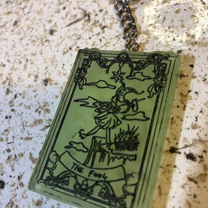 May include: A rectangular, light green keychain featuring a black illustration of a tarot card. The card depicts a figure on a cliff, with the words "THE FOOL" on a banner. The keychain has a silver chain and ring.
