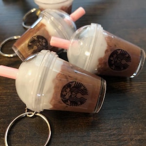May include: Miniature coffee drink keychains with a clear plastic cup, white whipped cream, and a pink straw. The Starbucks logo is visible on the cup. The keychains are on a dark wooden surface.