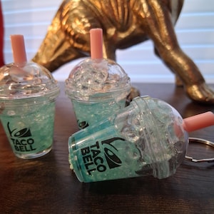 May include: Three clear plastic cups resembling bubble tea, filled with blue liquid and ice-like embellishments. Each cup has a pink straw and the Taco Bell logo. A golden dinosaur statue is in the background.