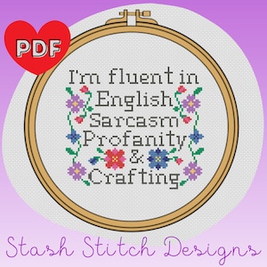 Peut inclure: Un motif de point de croix dans un cercle de broderie en bois avec le texte "Je suis fluide en anglais, sarcasme, grossièretés et artisanat". Le motif est entouré de fleurs colorées.