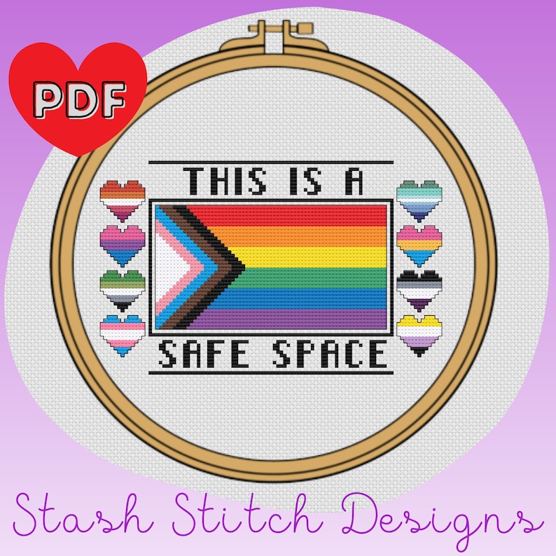 May include: A cross-stitch pattern featuring a rainbow pride flag with a black and brown chevron on the left side. The text "This is a Safe Space" is written below the flag. The pattern is surrounded by a wooden embroidery hoop. The pattern is available as a PDF download.
