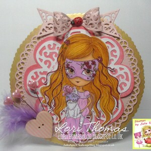 May include: A pink and gold card with a girl wearing a pink mask and holding a white cat. The card has a pink and white flower design and the text "Lori Thomas LoriThomasBlog.Blogspot.co.uk Proud Designer and Co-ordinator JuliaSpiriChallengeBlog.blogspot.co.uk Stamps by Julia Spiri" is printed on the card.