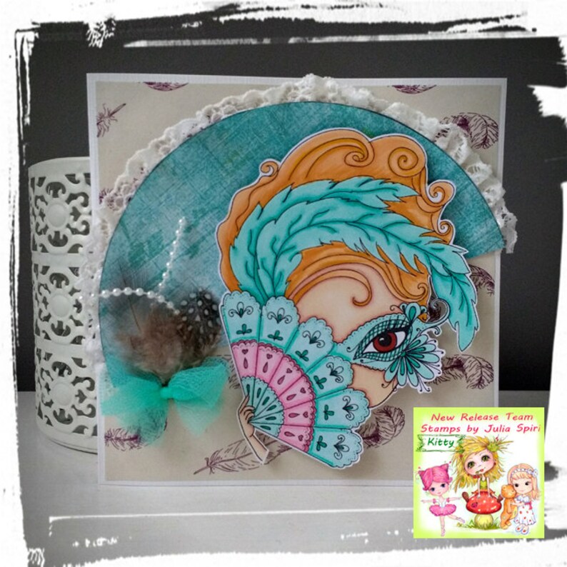 May include: A whimsical illustration of a girl with orange hair wearing a teal feather mask and holding a pink and teal fan. The image is on a card with a lace border and the text "New Release Team Stamps by Julia Spirit Kitty".