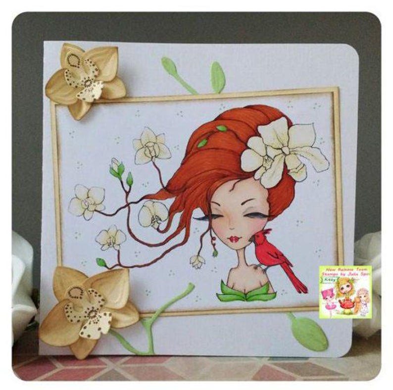 May include: A handmade greeting card featuring a whimsical illustration of a woman with red hair and a cardinal bird. The woman is surrounded by white flowers and green leaves. The card has a white background and a wooden frame.