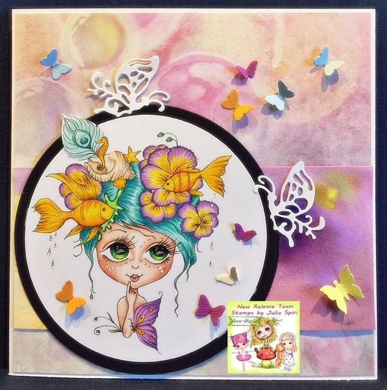 May include: A whimsical illustration of a girl with blue hair and large eyes. She is surrounded by flowers, fish, and a seashell. The illustration is framed in a black circle and surrounded by butterflies. The text "New Release Team Stamps by Julia Spirit" is visible in the lower right corner.